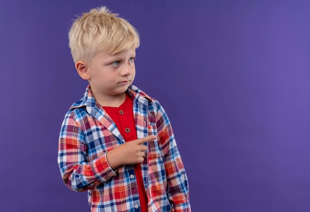 A cute little boy with blonde hair wearing checked shirt pointing with index finger at something on a purple wall