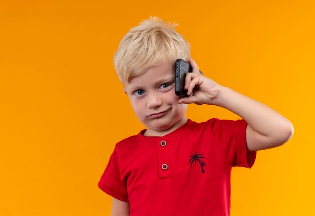 A cute little boy with blonde hair wearing red shirt holding mobile phone while looking on an orange wall