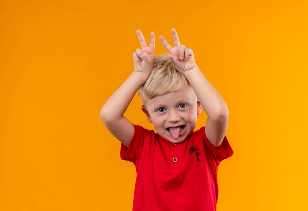 黄色い壁に頭の上に2本の指を置いた赤いTシャツを着たブロンドの髪のかわいい男の子