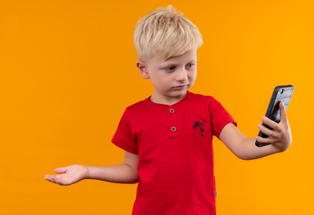 A cute little boy with blonde hair wearing red t-shirt looking at mobile phone with open hand on a yellow wall