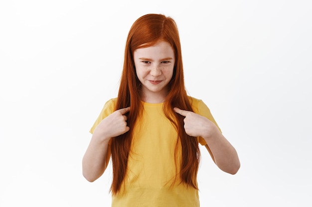 Cute little ginger girl with freckles points at herself with sly smile, choose me gesture, self-promoting, bragging, standing in yellow t-shirt against white background