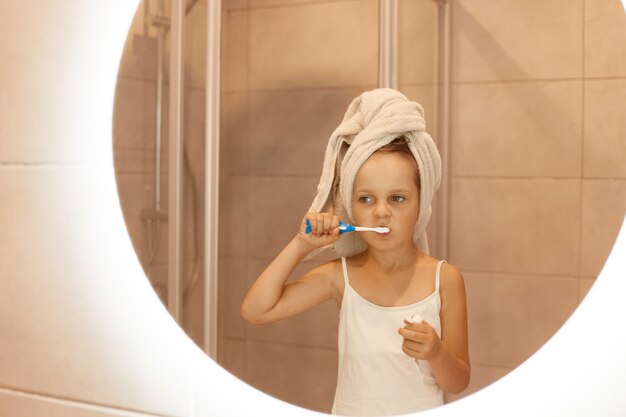 Cute little girl brushing teeth in bathroom while standing in front of mirror, wearing white sleeveless t shirt and wrapped her hair in towel, morning hygienic procedures.