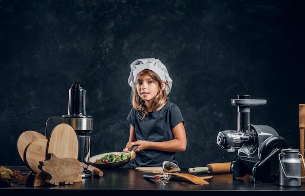 Милая маленькая девочка в шляпе шеф-повара готовит овощи для приготовления пищи на темном фоне.