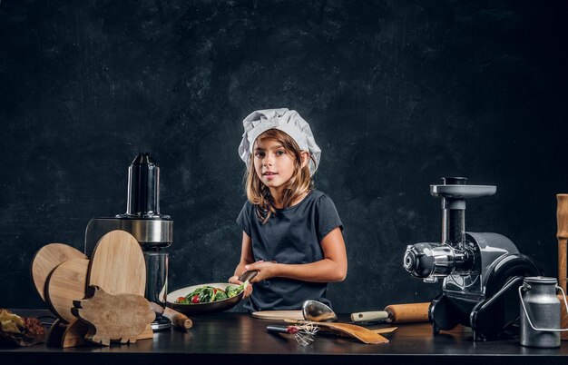 シェフの帽子をかぶったかわいい女の子は、暗い背景で調理するための野菜を準備しています。