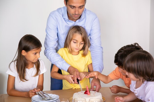 父の助けを借りてバースデーケーキを切るかわいい女の子。一緒に誕生日を祝い、ダイニングルームでデザートを待っている幸せな愛らしい子供たち。子供の頃、お祝い、休日の概念