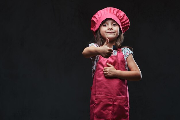 스튜디오에서 엄지손가락을 들고 포즈를 취하는 분홍색 요리사 옷을 입은 귀여운 소녀. 어두운 질감된 배경에 고립.