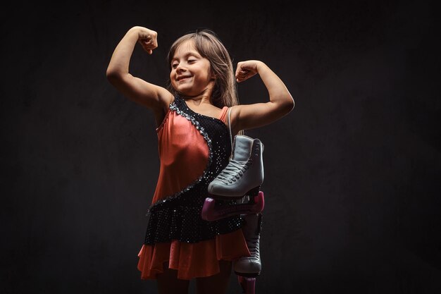 Cute little girl dressed in skater dress holds ice skates and shows muscles. Isolated on a dark textured background.