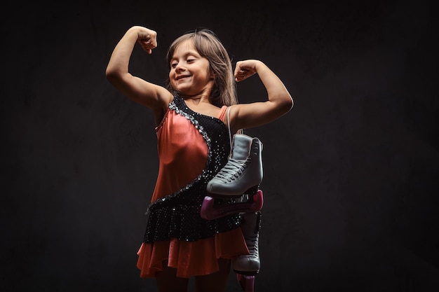 Cute little girl dressed in skater dress holds ice skates and shows muscles. Isolated on a dark textured background.