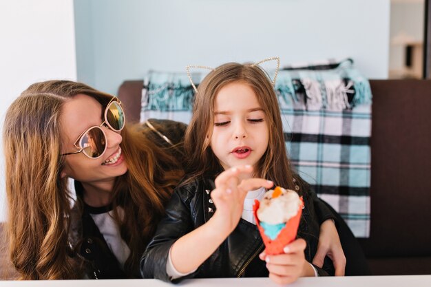 Cute little girl and her attractive cheerful mom wearing trendy sunglasses having fun at home and eating dessert.