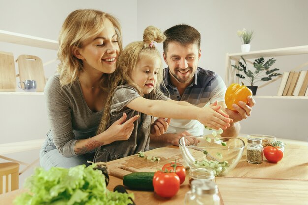 Cute little girl and her beautiful parents are cutting vegetables and smiling while making salad in kitchen at home. Family lifestyle concept
