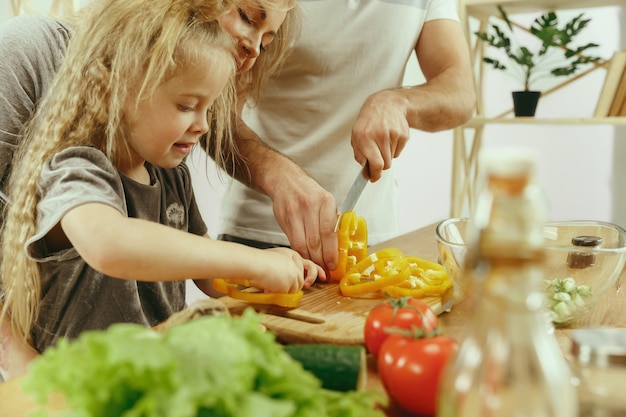 Cute little girl and her beautiful parents are cutting vegetables and smiling while making salad in kitchen at home. Family lifestyle concept