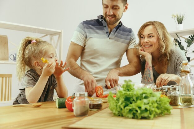 Cute little girl and her beautiful parents are cutting vegetables and smiling while making salad in kitchen at home. Family lifestyle concept