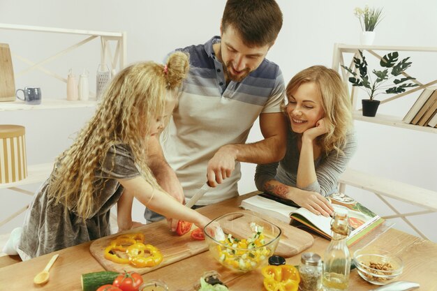 Foto gratuita la bambina sveglia ei suoi bellissimi genitori stanno tagliando le verdure e sorridendo mentre fanno l'insalata in cucina a casa. concetto di stile di vita familiare