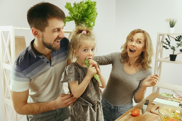 귀여운 소녀와 그녀의 아름다운 부모는 야채를 자르고 집에서 부엌에서 샐러드를 만드는 동안 웃고 있습니다. 가족 라이프 스타일 개념