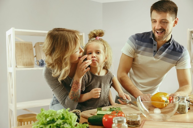 La bambina sveglia ei suoi bellissimi genitori stanno tagliando le verdure e sorridendo mentre fanno l'insalata in cucina a casa. concetto di stile di vita familiare