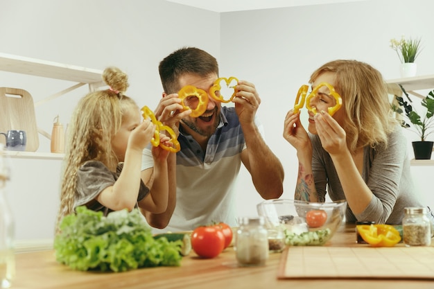 La bambina sveglia ei suoi bellissimi genitori stanno tagliando le verdure e sorridendo mentre fanno l'insalata in cucina a casa