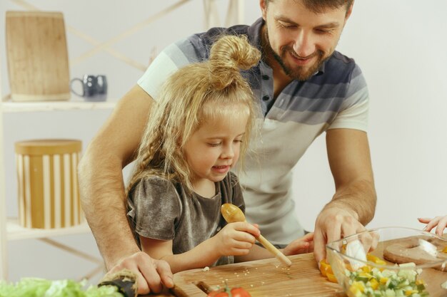 Cute little girl and her beautiful parents are cutting vegetables and smiling while making salad in kitchen at home