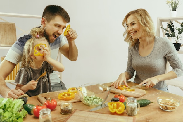 Cute little girl and her beautiful parents are cutting vegetables and smiling while making salad