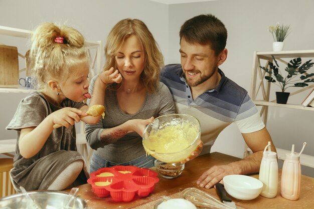 Милая маленькая девочка и ее красивые родители готовят тесто для торта на кухне дома