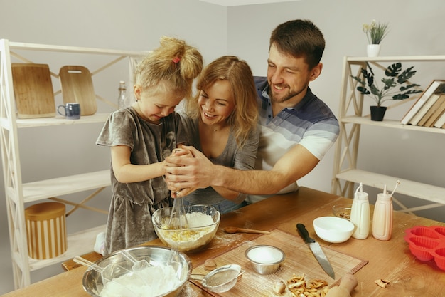 Bambina sveglia ed i suoi bei genitori che preparano la pasta per la torta in cucina a casa.