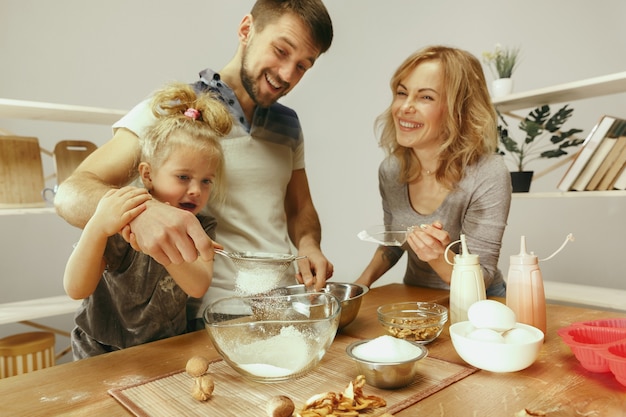 Cute little girl and her beautiful parents preparing the dough for the cake in kitchen at home.