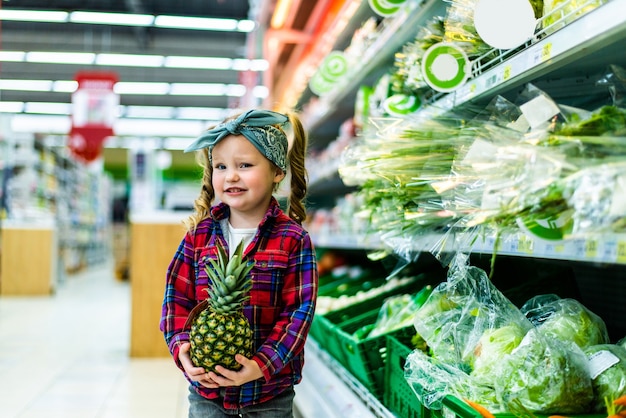 Cute little girl holding a pineapple in a food store or supermarket