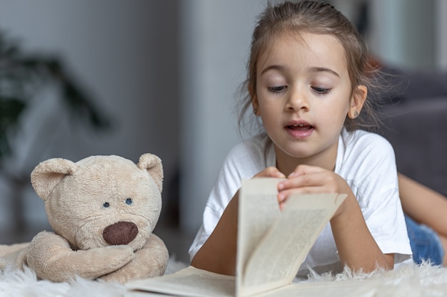 Cute little girl at home, lying on the floor with her favorite toy and reads book.