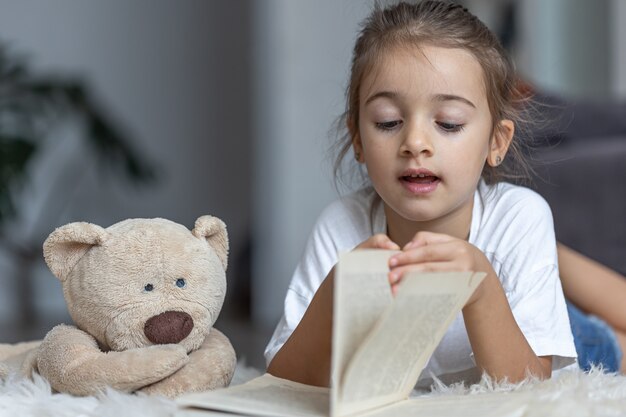 집에 있는 귀여운 소녀가 좋아하는 장난감을 들고 바닥에 누워 책을 읽습니다.