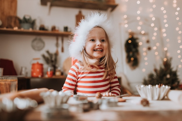 산타 모자를 쓴 귀여운 소녀가 크리스마스 테이블 겨울 새해 부엌에서 장난을 치고 있다 프리미엄 사진