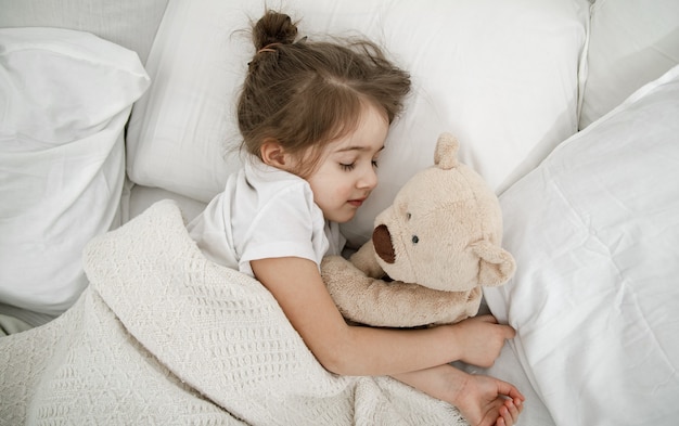 귀여운 소녀가 테디 베어 장난감을 가지고 침대에서 자고 있습니다. 아동 발달과 수면의 개념. 상단에서보기.