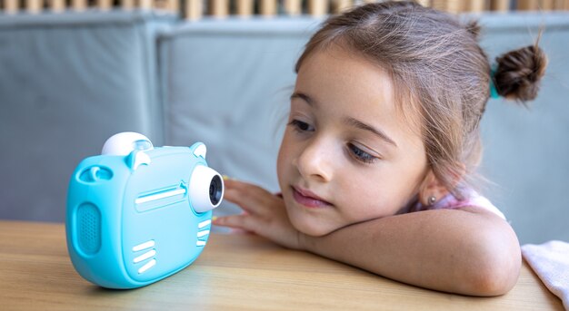 귀여운 소녀가 즉석 사진 인화를 위해 아이들의 파란색 장난감 카메라를 쳐다보고 있습니다.