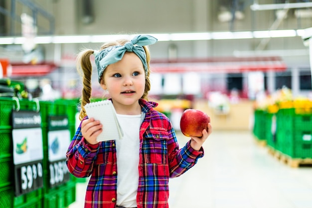Милая маленькая девочка делает список товаров для покупки в супермаркете