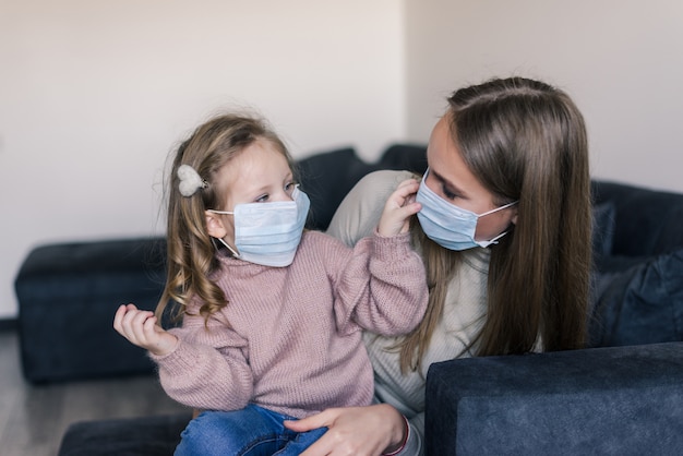 Cute little girl and mother wearing face mask, sitting on bed at home, consoling sad preschool daughter