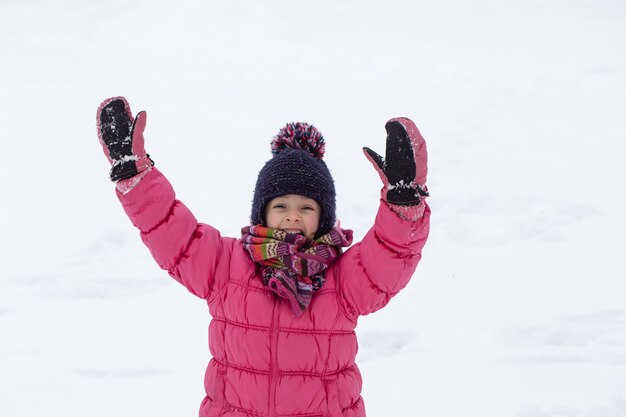 A cute little girl in a pink jacket and a hat is playing in the snow. Winter children's entertainment concept.