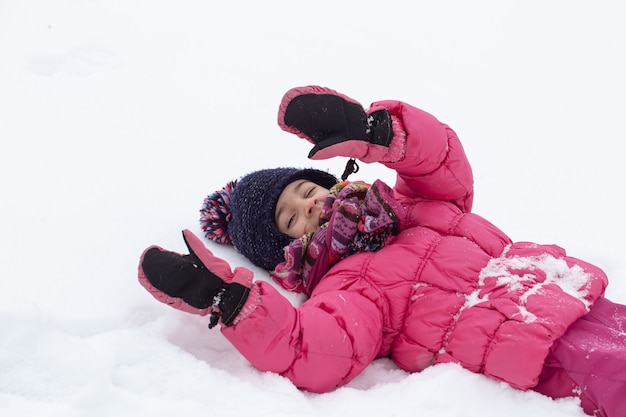 A cute little girl in a pink jacket and a hat is playing in the snow. Winter children's entertainment concept.
