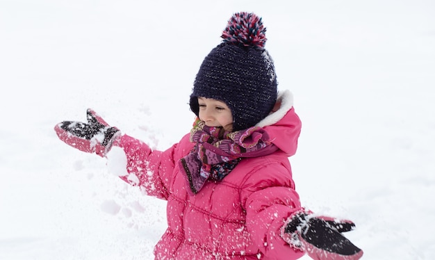 분홍색 재킷과 모자를 입은 귀여운 소녀가 눈 속에서 놀고 있습니다. 겨울 어린이 엔터테인먼트 개념.