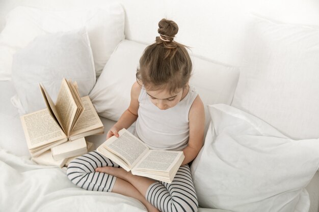 Cute little girl reading a book on the bed in the bedroom.