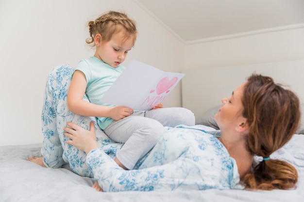 Cute little girl reading greeting card while sitting on mother's stomach at home