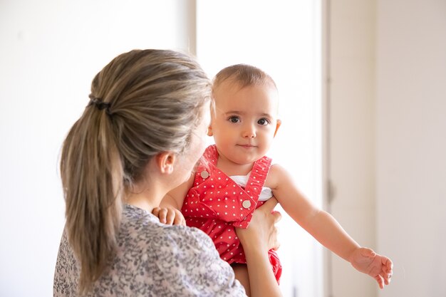 Cute little girl in red suit sitting on mother hands. Back view of caring mother holding daughter and talking with her. Family time, motherhood and being at home concept