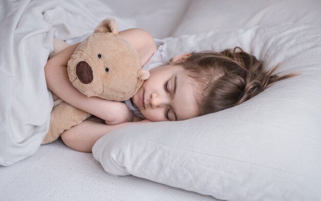 귀여운 소녀는 부드러운 곰 장난감, 어린이 휴식과 수면의 개념이있는 흰색 아늑한 침대에서 달콤하게 잔다.