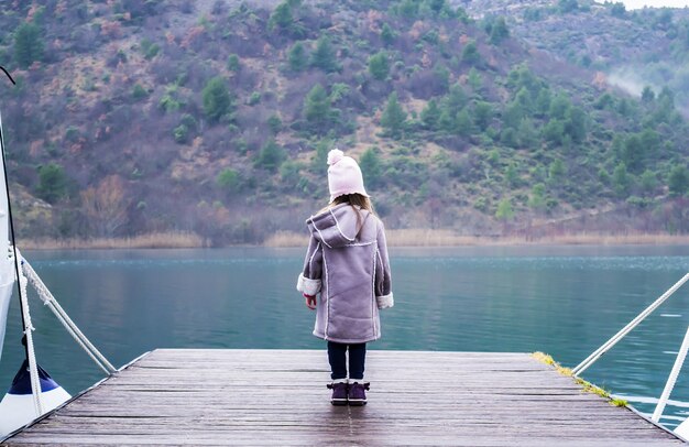 Cute little girl standing on the wooden pier near the turquoise lake