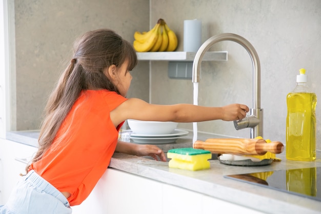 Foto gratuita bambina sveglia che lava il piatto in cucina da sola. bambino che raggiunge il rubinetto del rubinetto del lavello della cucina e accende l'acqua.