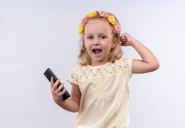 Милая маленькая девочка в желтой рубашке в цветочной повязке на голову держит руку на голове с открытым ртом, держа мобильный телефон на белой стене