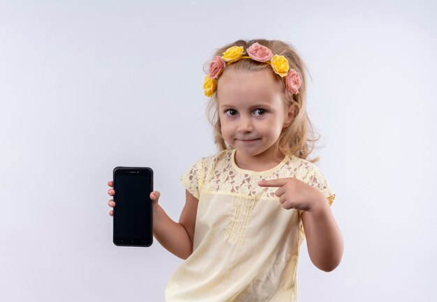 A cute little girl wearing yellow shirt in floral headband pointing at blank space of mobile phone with index finger on a white wall