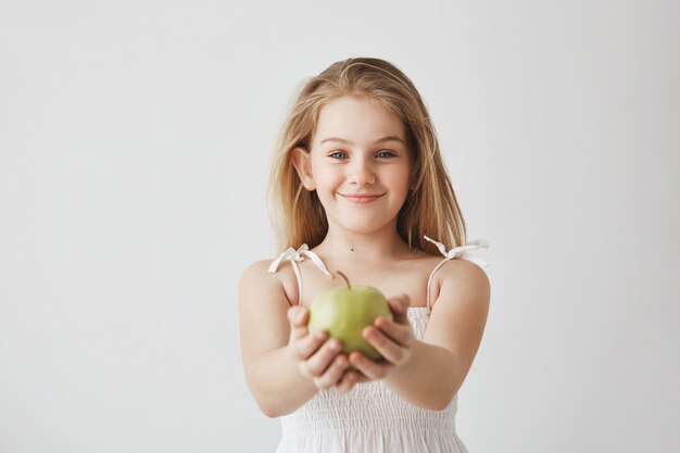 Cute little girl with blond long hair and blue eyes in white dress smiling, holding apple in hands and showing