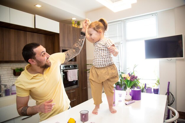 Cute little girl with her happy cheerful father holding his arm standing on the kitchen table trying to make a step having fun together in the kitchen at home happy family concept