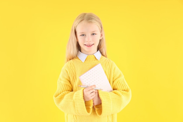 Cute little girl with notebook and pen on yellow background