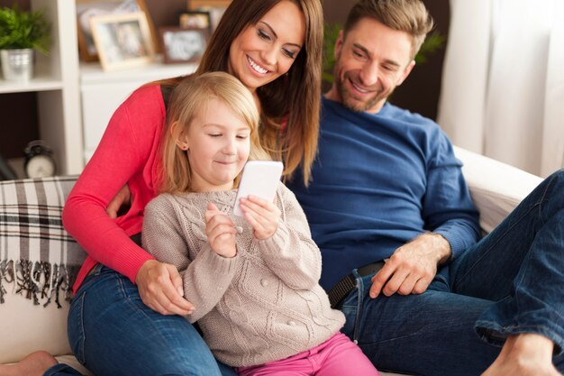 Cute little girl with parents playing by mobile phone at home