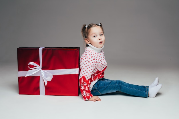 Милая маленькая девочка с хвостиками в зимнем свитере и джинсах сидит спиной к красному обернутому рождественскому подарку с белым бантом. Ребенок надувает губы на камеру.