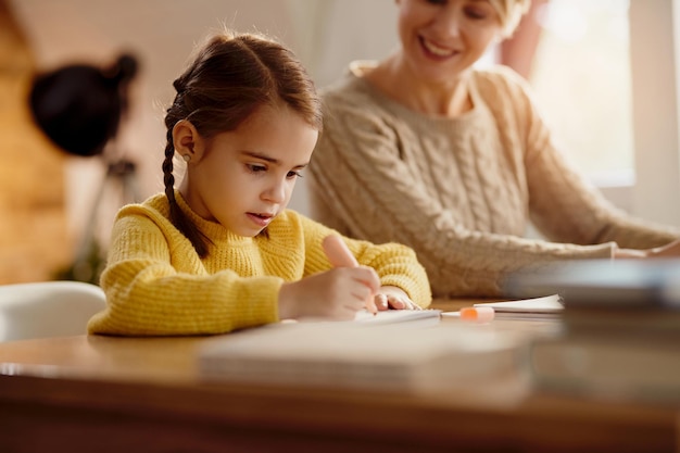 Cute little girl writing while her mother is working beside her at home